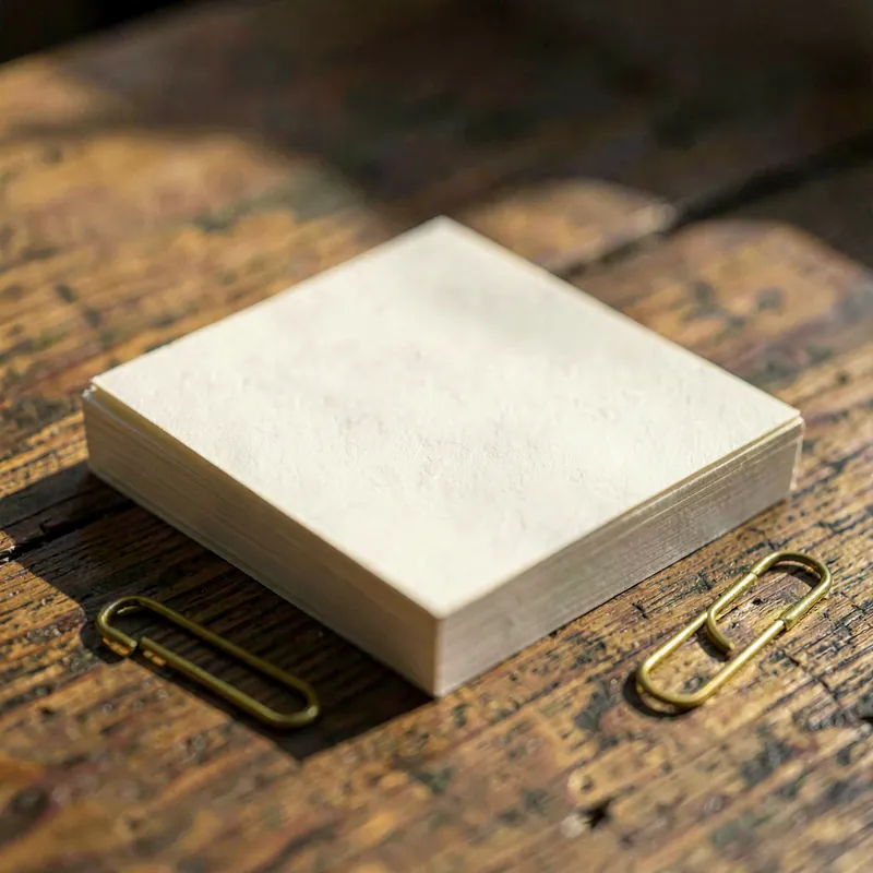 A neat squared stack of blank cream paper on a timber table beside a plain brass paperclip